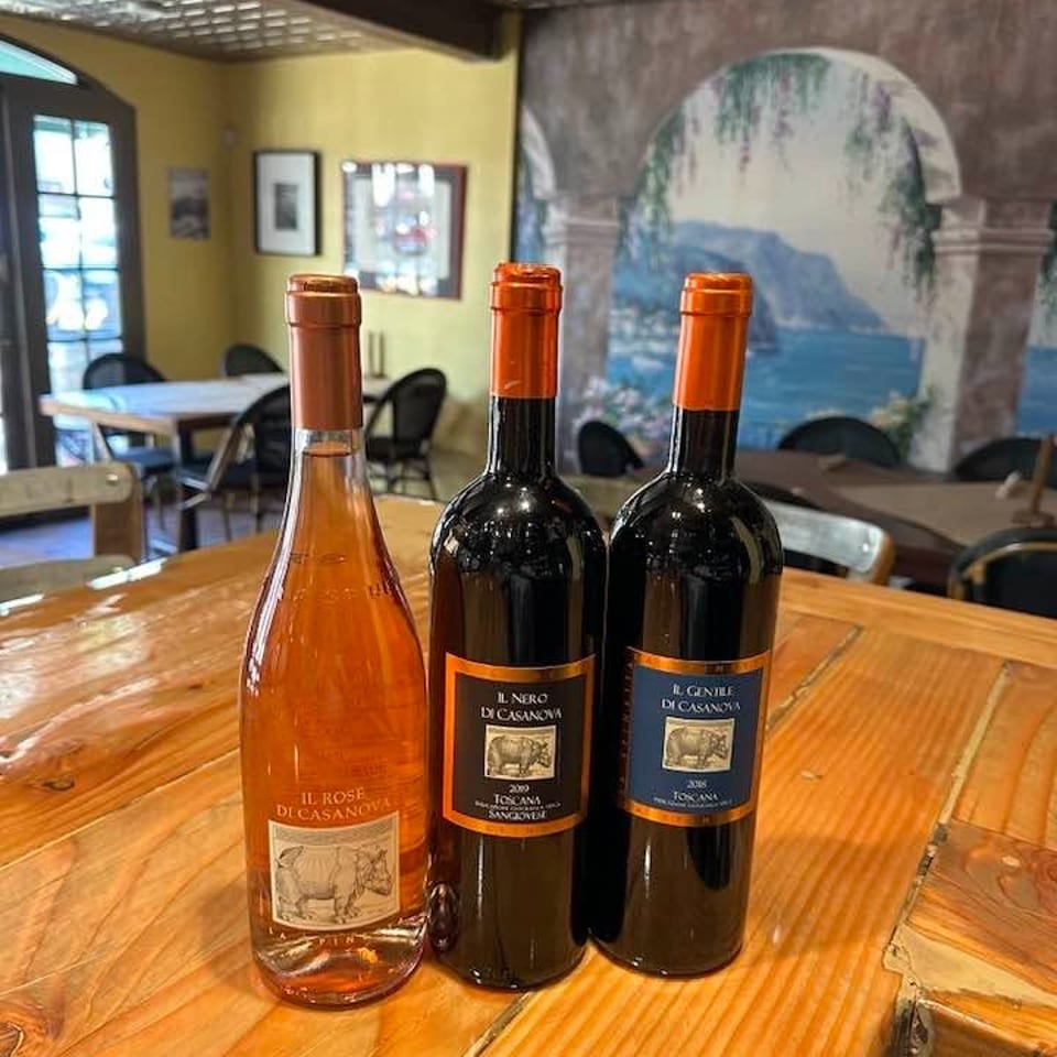 Three wine bottles displayed on a wooden restaurant table, with a rustic dining room and scenic mural in the background.
