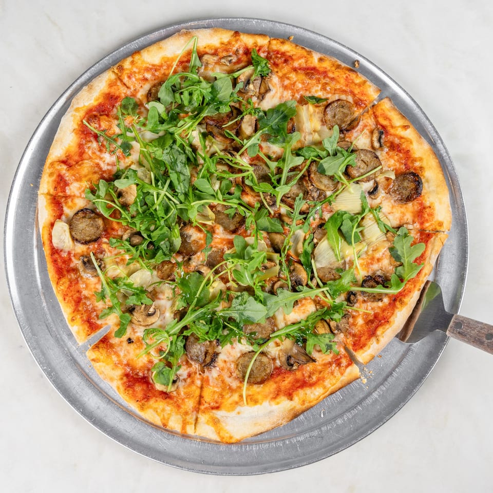 Thin-crust pizza topped with melted cheese, mushrooms, artichokes, sausage, and fresh arugula on a metal serving tray.