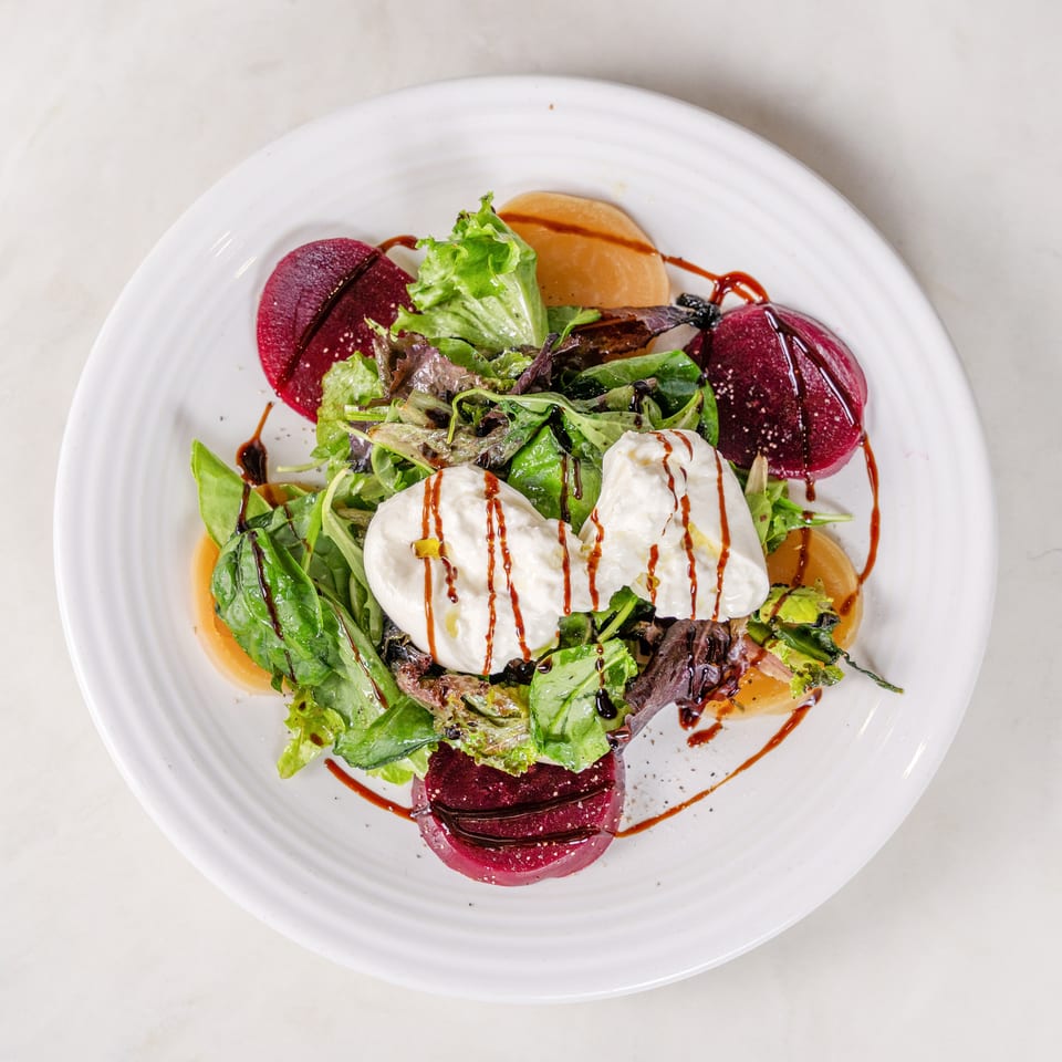 Mixed green salad with burrata, sliced beets, peaches, and balsamic glaze on a white plate.
