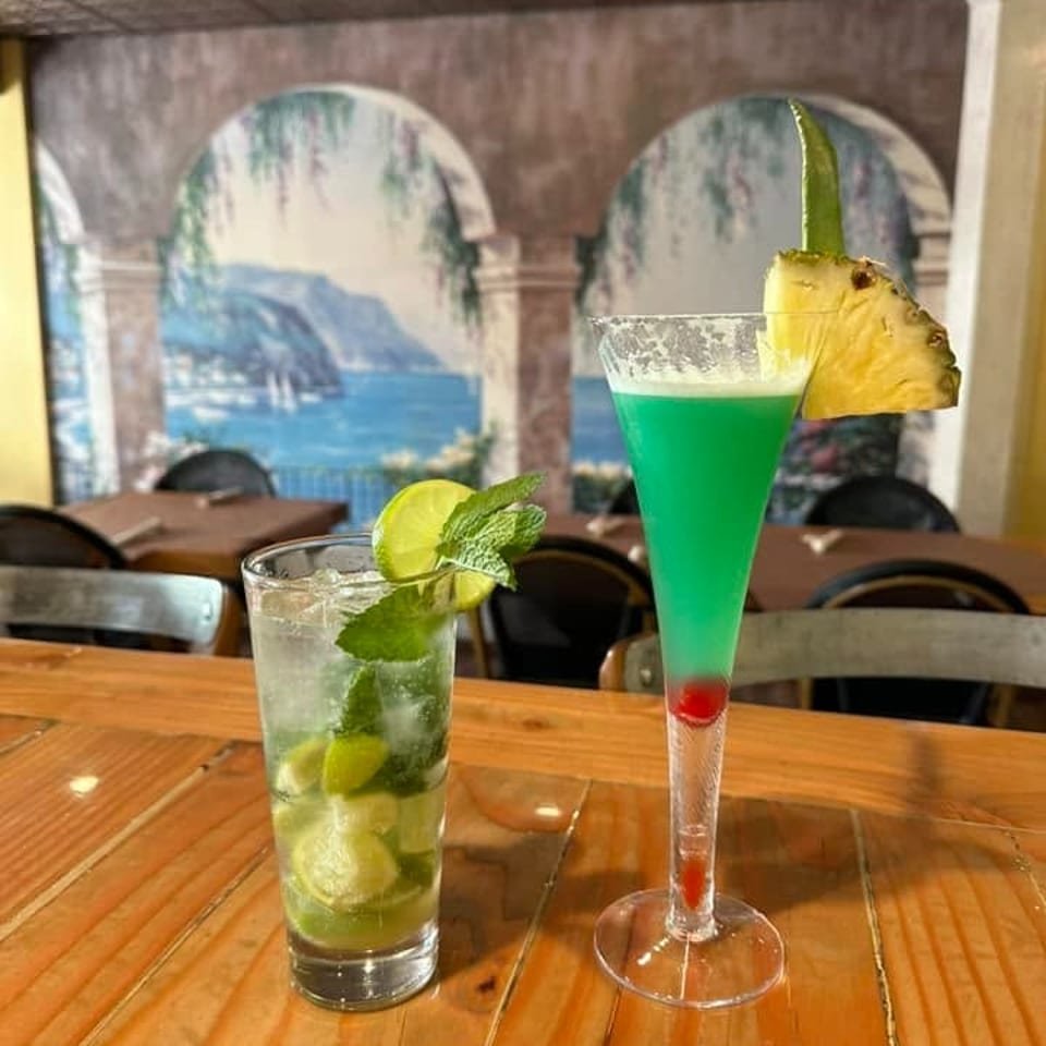Two colorful cocktails sit on a wooden restaurant table, including a mint-and-lime drink and a pineapple-garnished layered green cocktail.