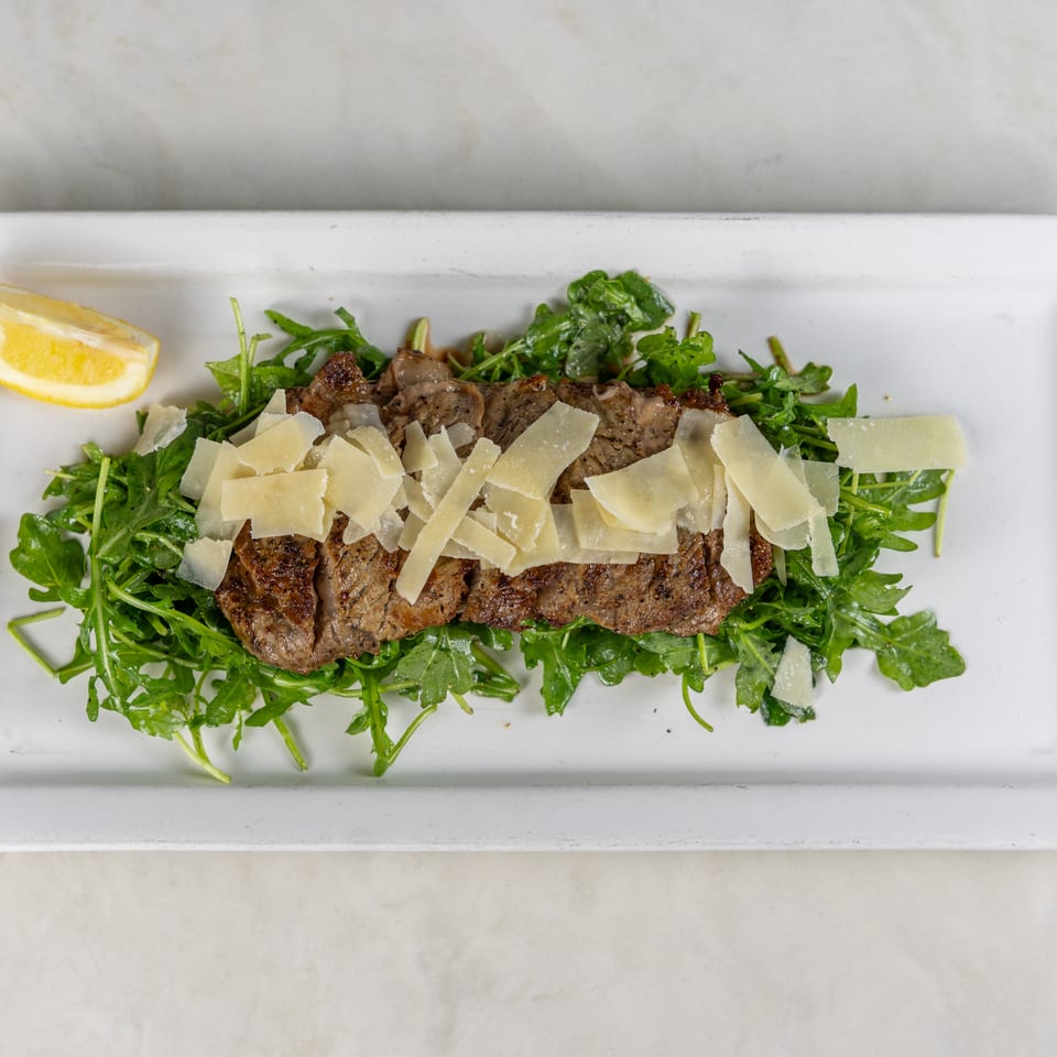 Grilled steak slices topped with shaved Parmesan on arugula, served with a lemon wedge on a white platter.