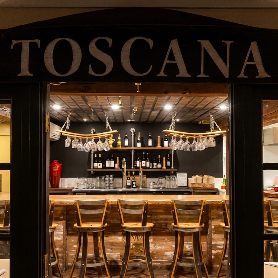 TOSCANA restaurant entrance revealing a warmly lit rustic bar with hanging wine glasses, bottles, and wooden bar stools.