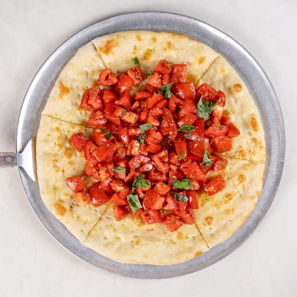 Sliced flatbread topped with chopped tomatoes, fresh basil, olive oil, and herbs on a metal serving tray.