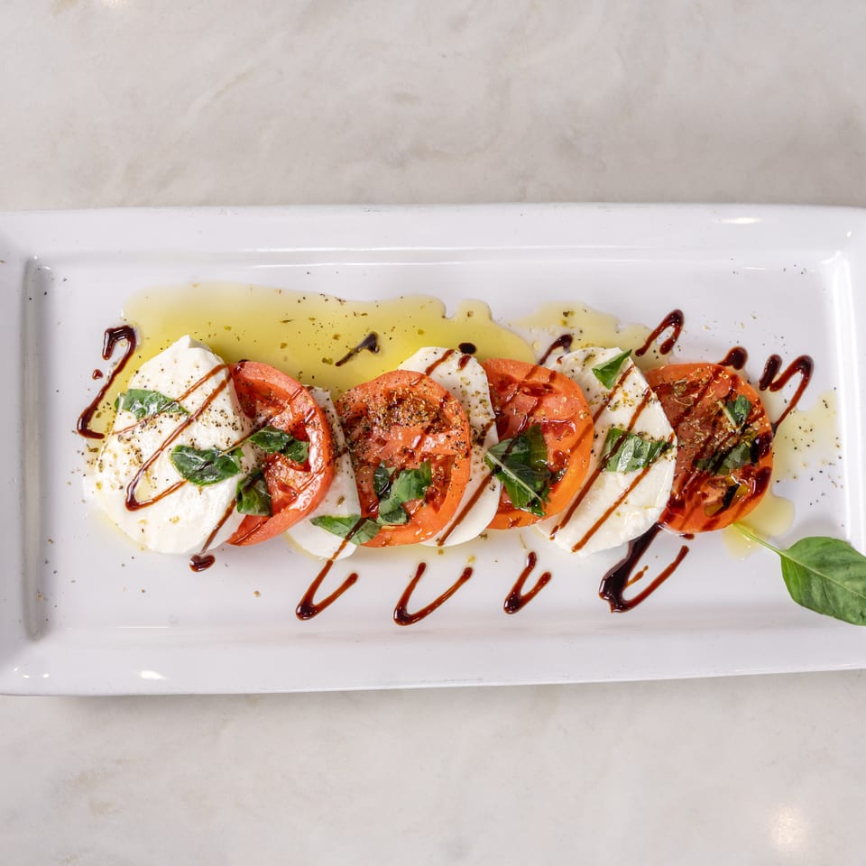 Caprese salad with sliced tomatoes, fresh mozzarella, basil, olive oil, and balsamic drizzle on a rectangular white plate.