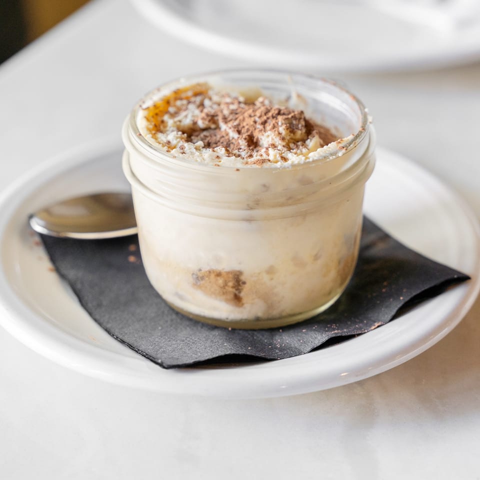 Individual tiramisu dessert in a glass jar, topped with whipped cream and cocoa, served on a black napkin with a spoon.