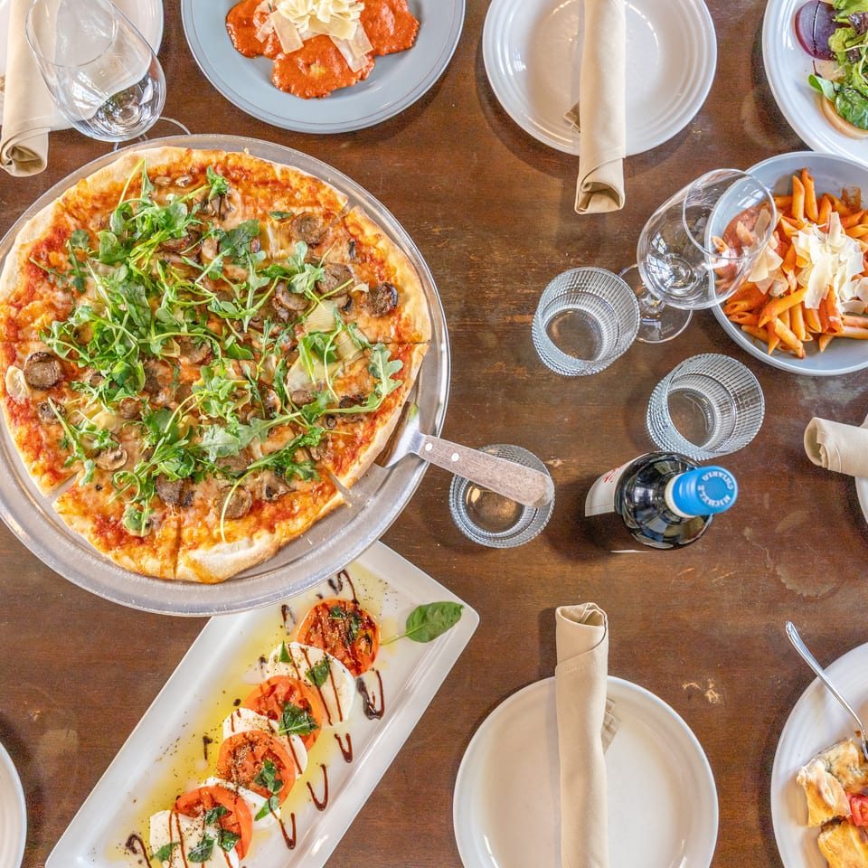 Overhead view of an Italian restaurant table with pizza, penne pasta, salad, caprese, wine glasses, and folded napkins.