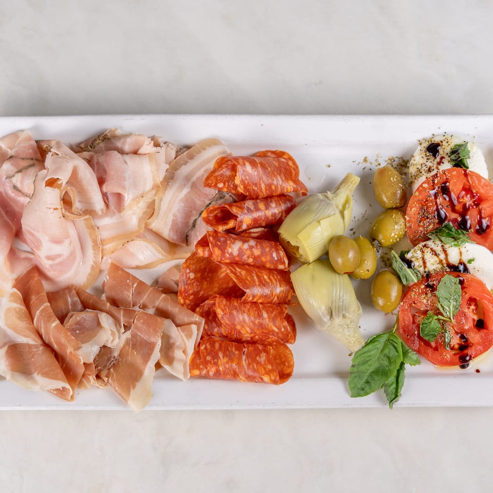 Antipasto platter with prosciutto, spicy salami, olives, artichokes, burrata, and tomatoes garnished with basil and balsamic glaze.