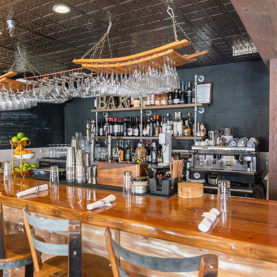 Rustic restaurant bar with hanging wine glasses, liquor shelves, espresso machine, polished wooden counter, and napkin-covered bar stools.