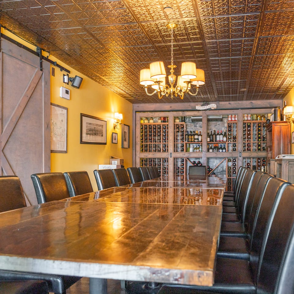 Elegant restaurant dining room with a long polished table, leather chairs, ornate tin ceiling, chandelier, and extensive wine display.