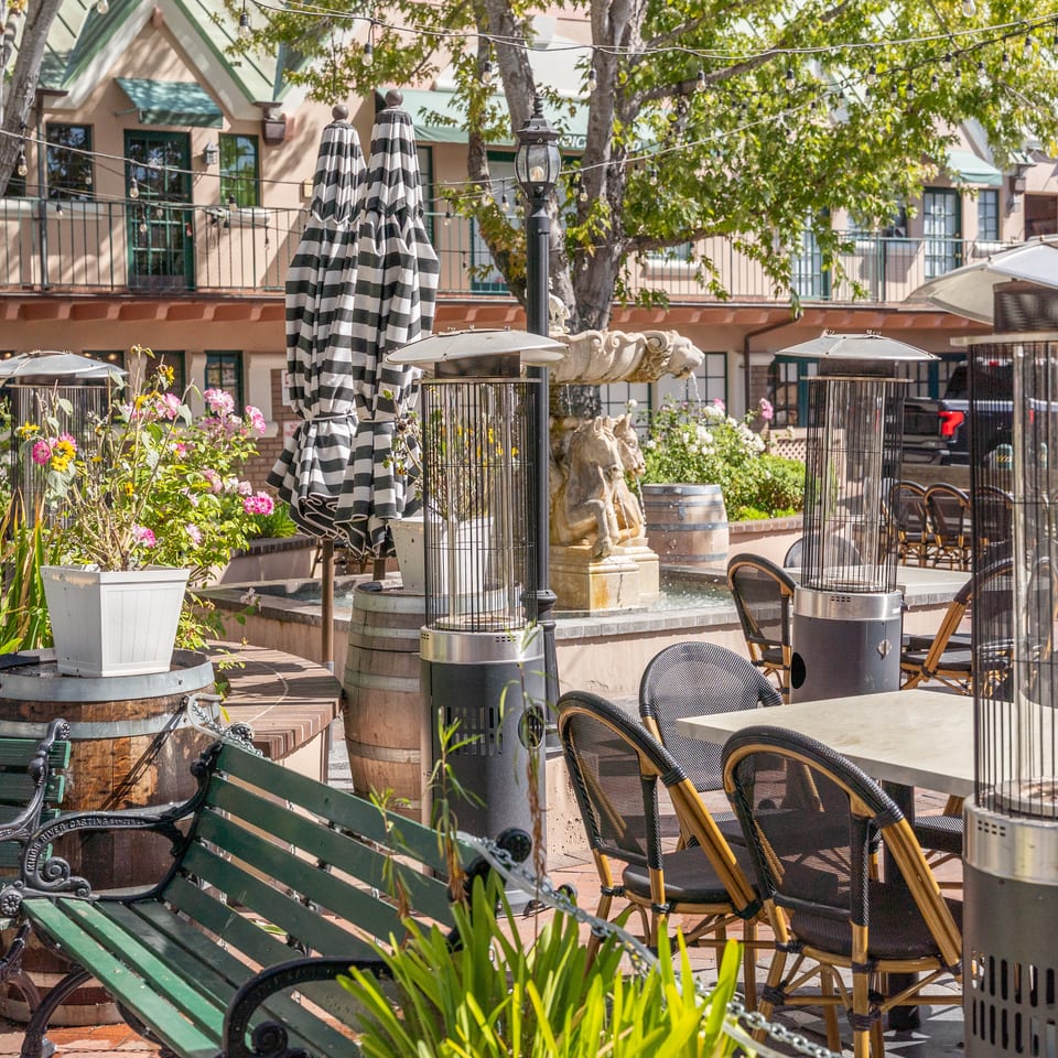 Outdoor restaurant patio with striped umbrellas, patio heaters, café tables, flowers, string lights, and a fountain.