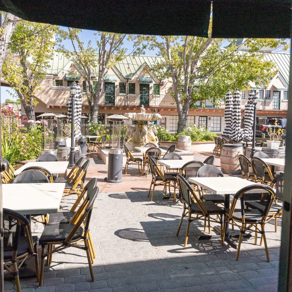Sunny restaurant patio with café tables, wicker chairs, striped umbrellas, string lights, trees, and a central fountain.