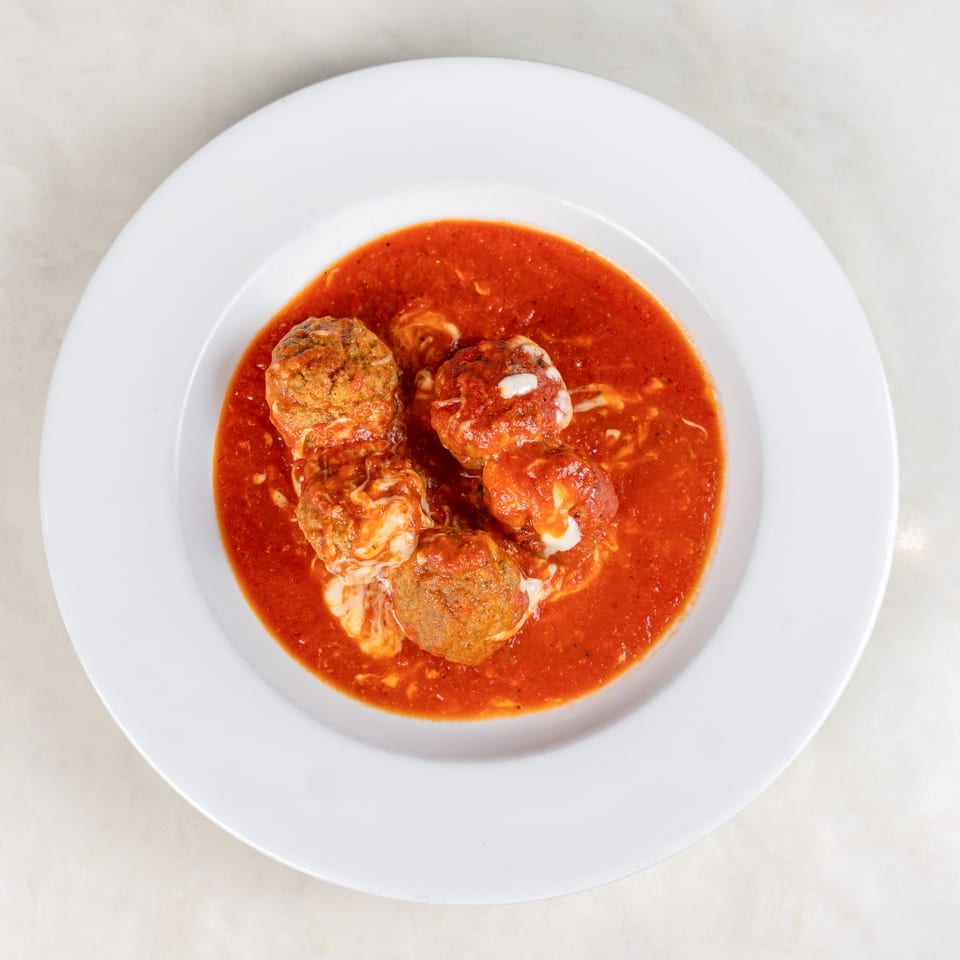 Five meatballs topped with melted cheese sit in bright tomato sauce on a white plate.