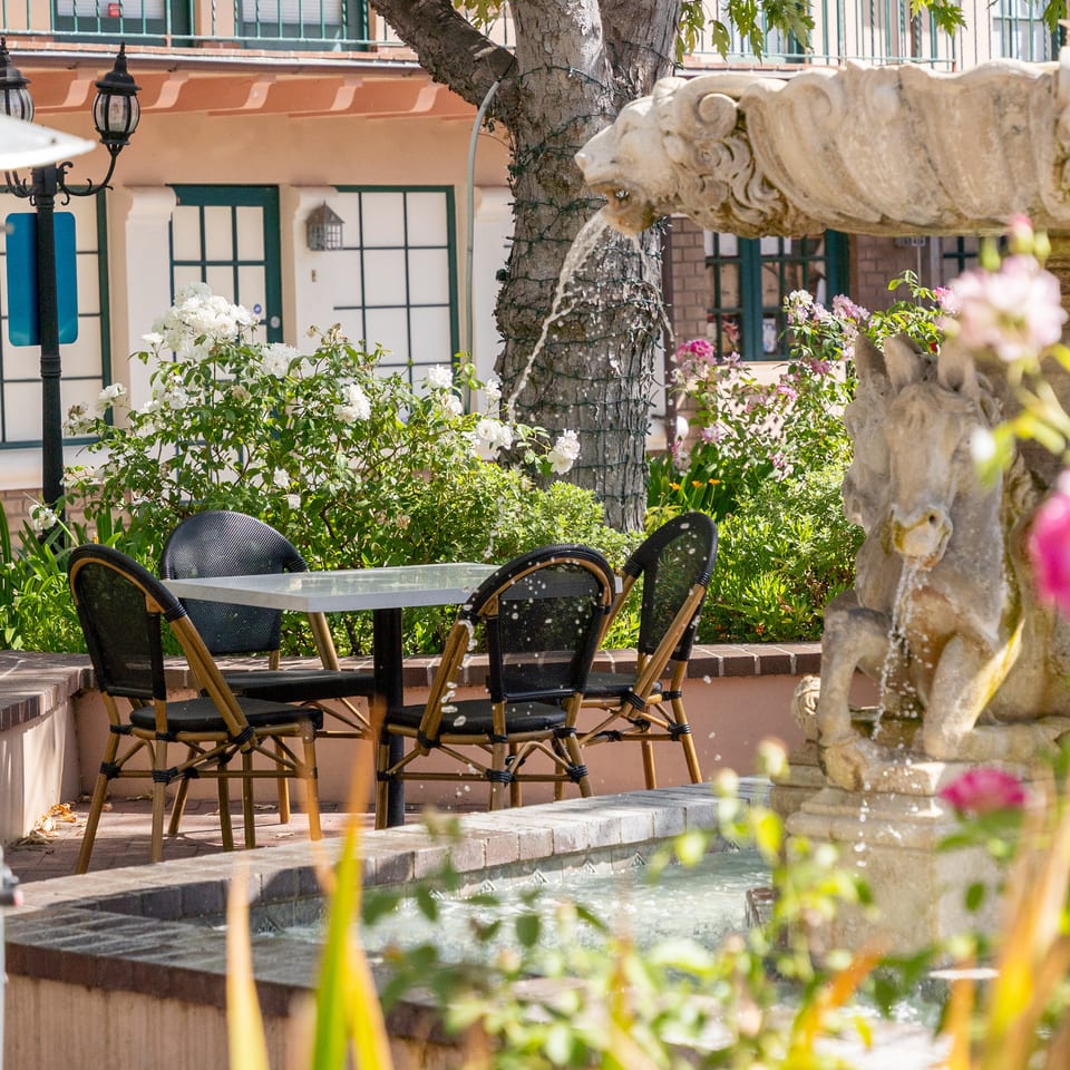 Outdoor restaurant patio with café chairs, flowering plants, a tree, and an ornate lion fountain.