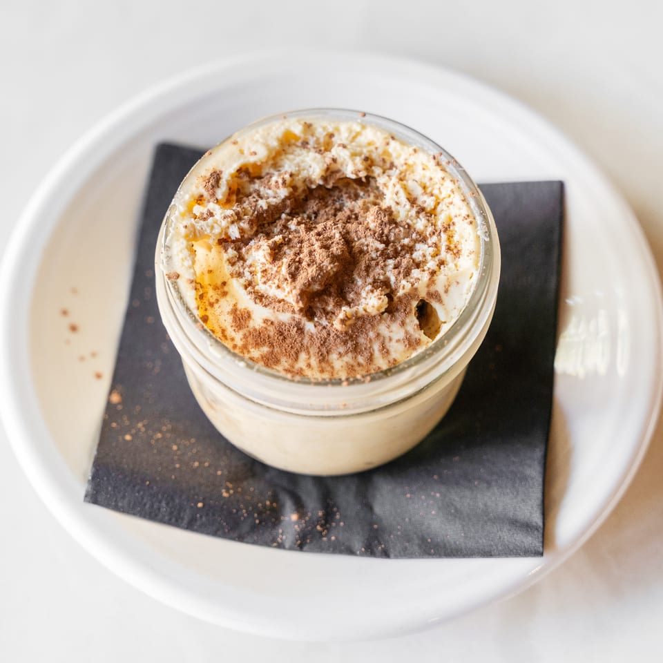 Creamy dessert topped with whipped cream and cocoa powder, served in a glass dish on a black napkin.