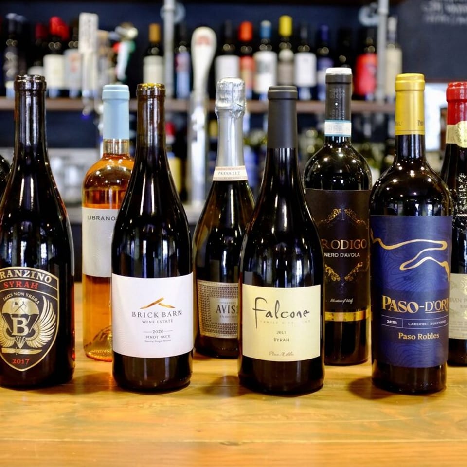 Assorted wine bottles displayed on a wooden bar, with shelves of additional bottles and a restaurant logo in the background.