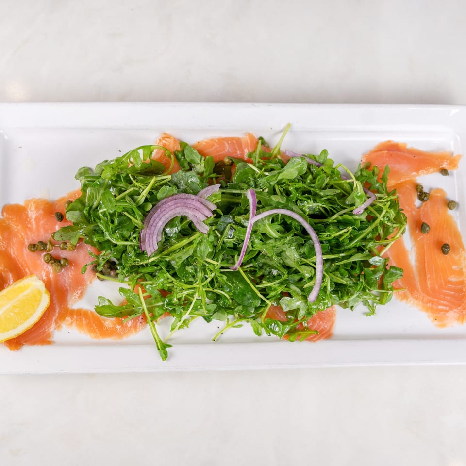 Smoked salmon slices surround an arugula salad topped with red onion, capers, and lemon wedges on a white platter.